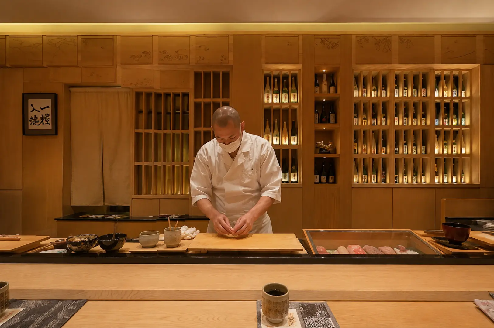 A sushi chef in white preparing food behind a traditional wooden counter, with shelves of sake bottles warmly illuminated in the background at Shinji by Kanesaka.