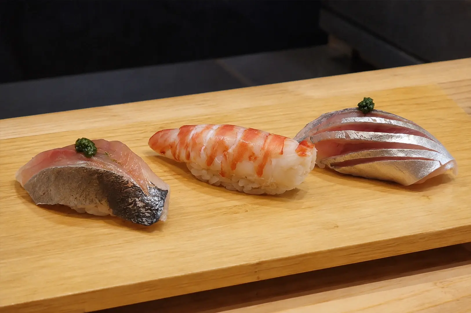Three pieces of carefully prepared nigiri on a wooden sushi board, featuring shrimp and silver-skinned fish, at Shinji by Kanesaka.