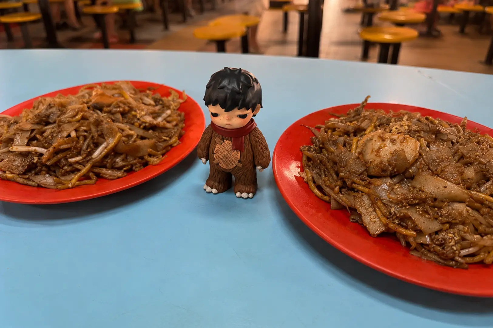 Two red plates of dark, wok-fried Singapore char kway teow with noodles, bean sprouts, meat and egg.