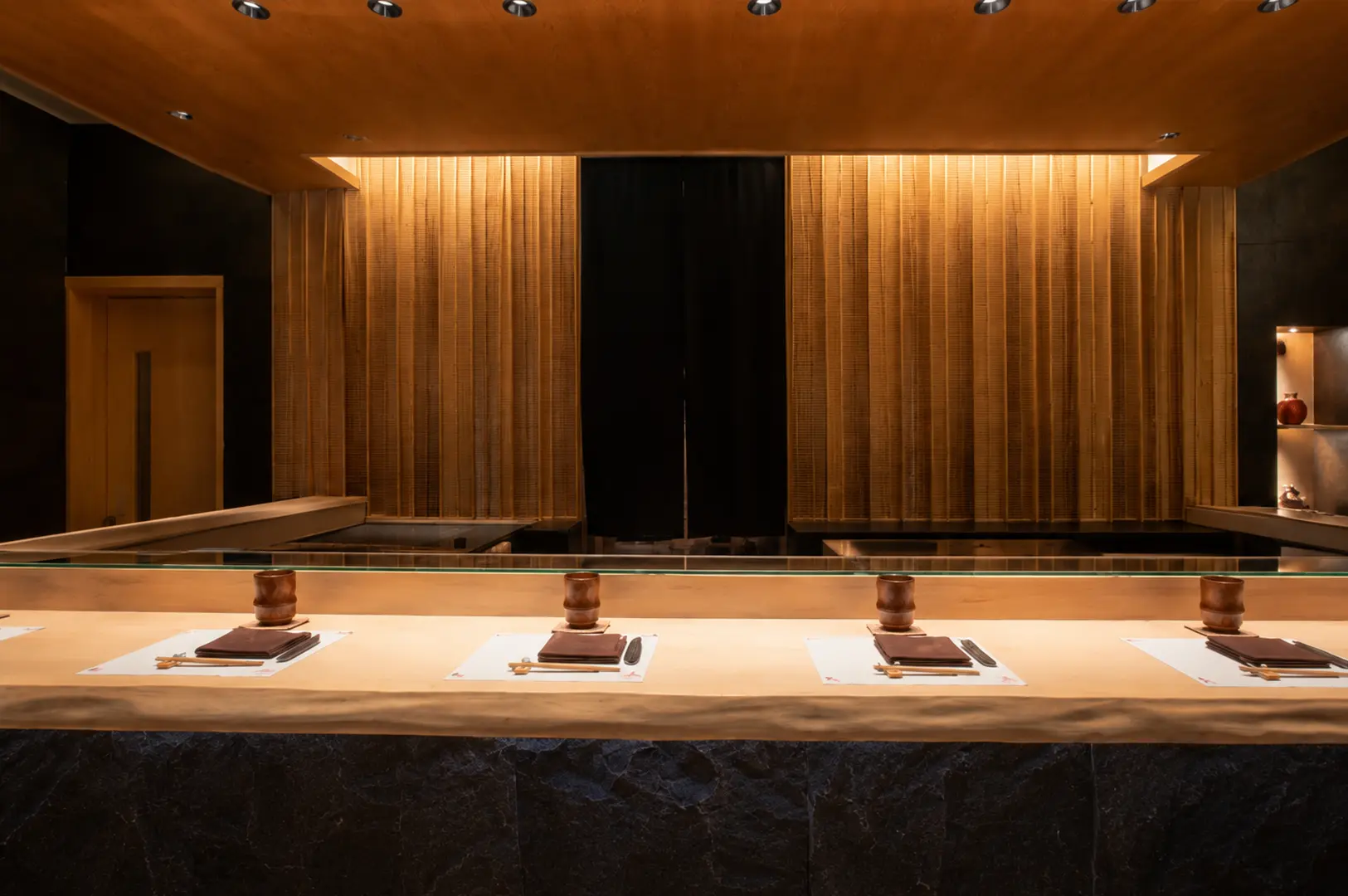 An elegant sushi counter with light wood finishes, neatly arranged place settings, and a warmly lit bamboo-textured wall at Sushi Kimura.