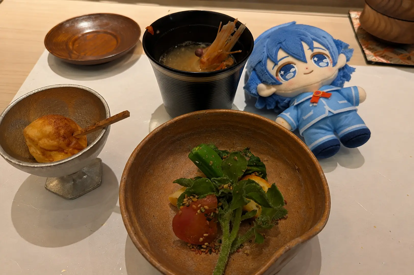 A selection of Japanese dishes on rustic ceramic plates, including vegetables and a small appetizer, with a blue anime plush beside the meal at Sushi Kimura.