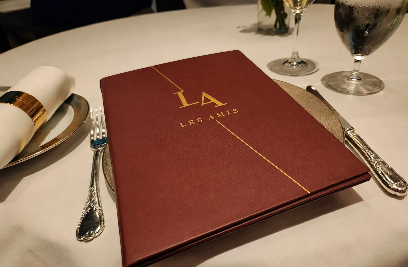 Les Amis menu cover displayed on a formally set fine dining table with silverware and wine glasses.