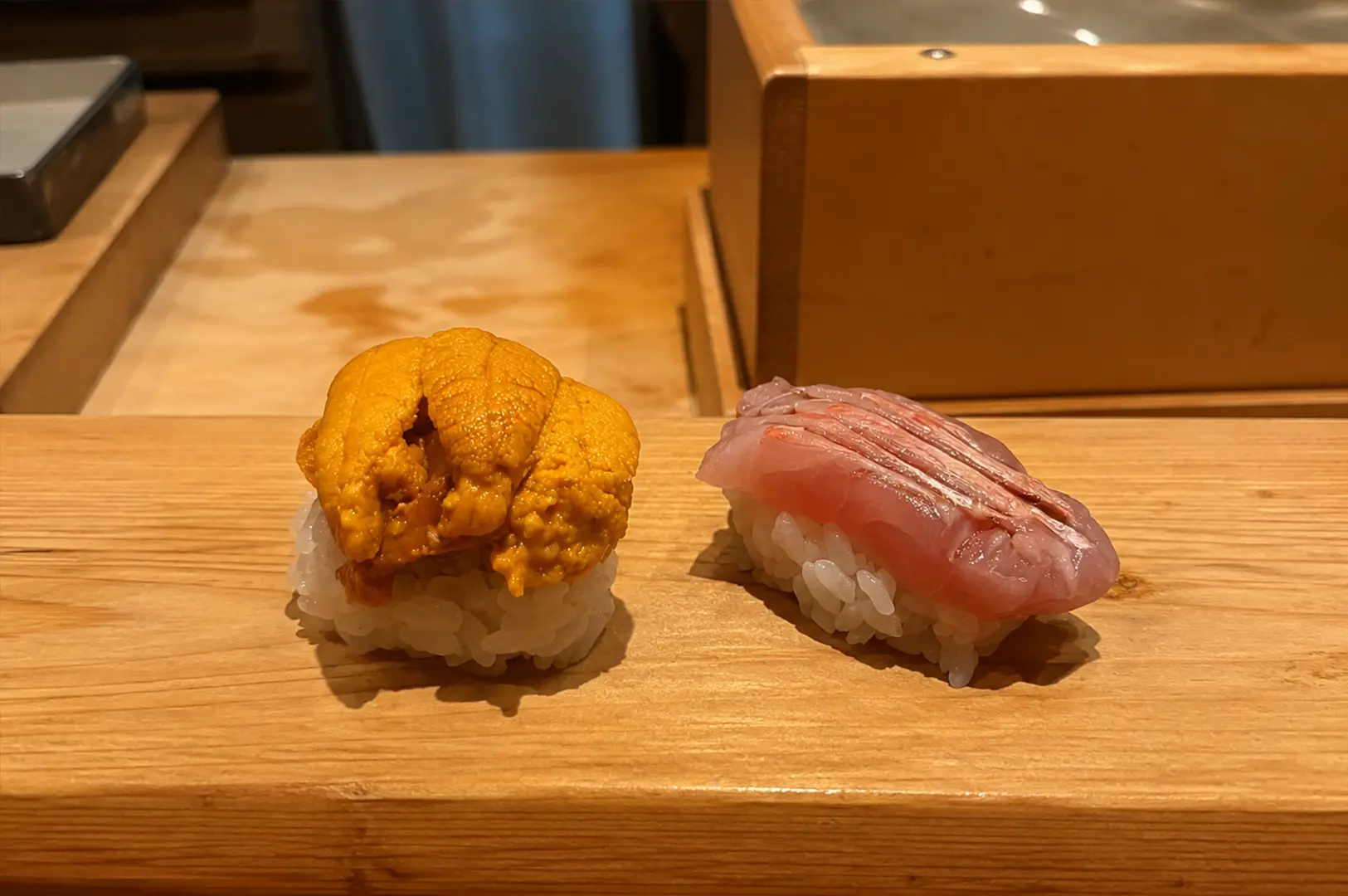 Two pieces of nigiri on a wooden board, one topped generously with sea urchin and the other with finely layered pink fish, at Sushi Ashino.