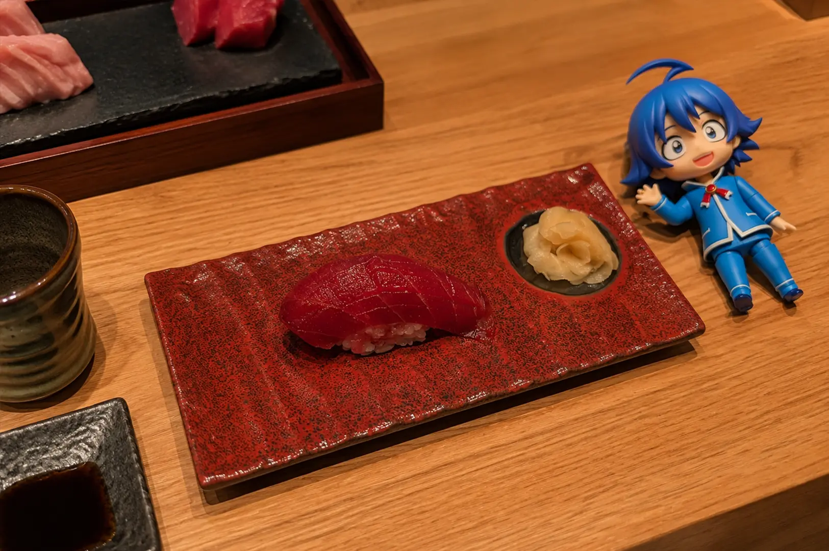A close-up of two pieces of sushi on a textured red ceramic plate: one topped with deep red tuna and the other with pale pink fish, accompanied by pickled ginger and soy sauce. A blue-haired anime figure sits beside the plate on a wooden table.