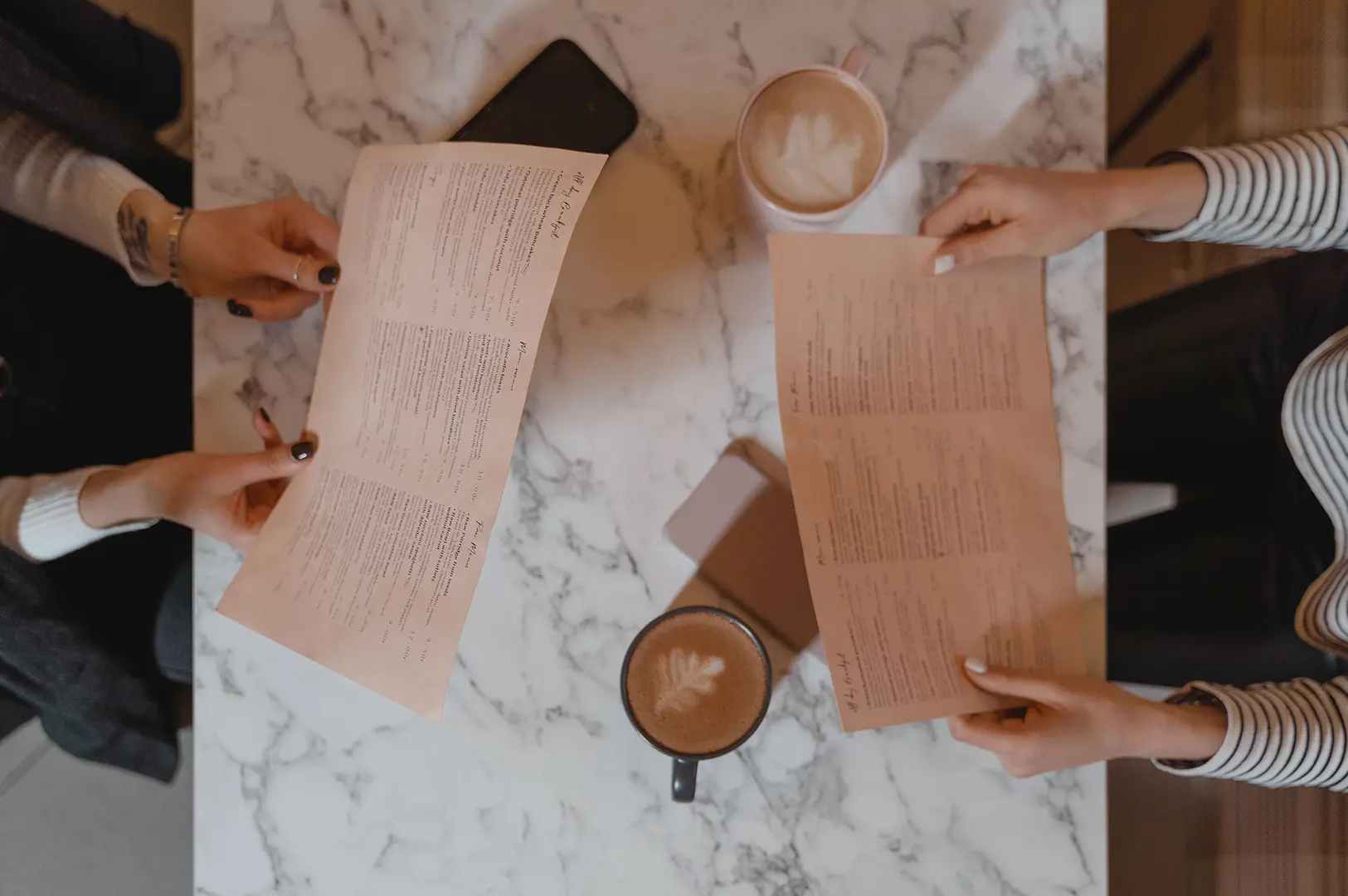 Two people looking over restaurant menus at a marble table with coffee drinks.