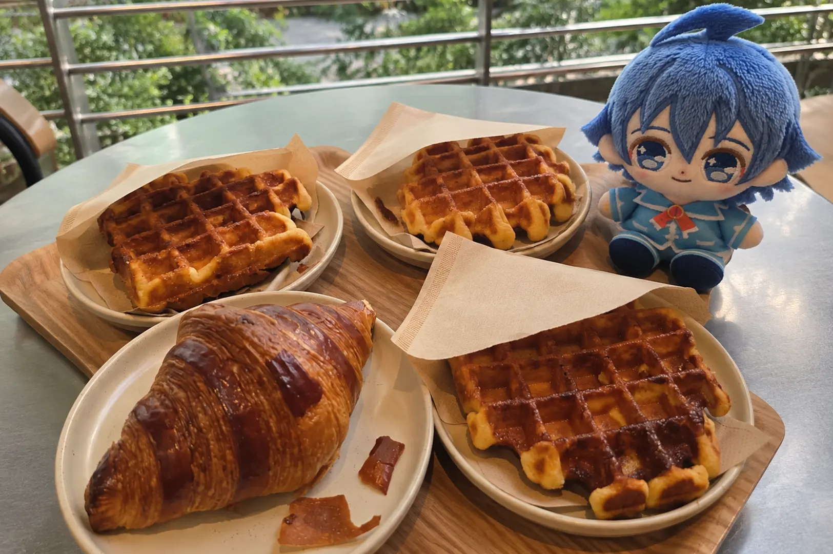 A café table filled with waffles and a croissant, with a blue plush toy sitting beside the food.