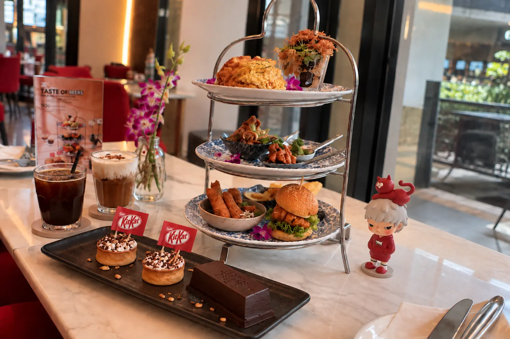 Three-tier afternoon tea stand with savory bites, desserts, KitKat treats, drinks, and a red character figurine on a café table.