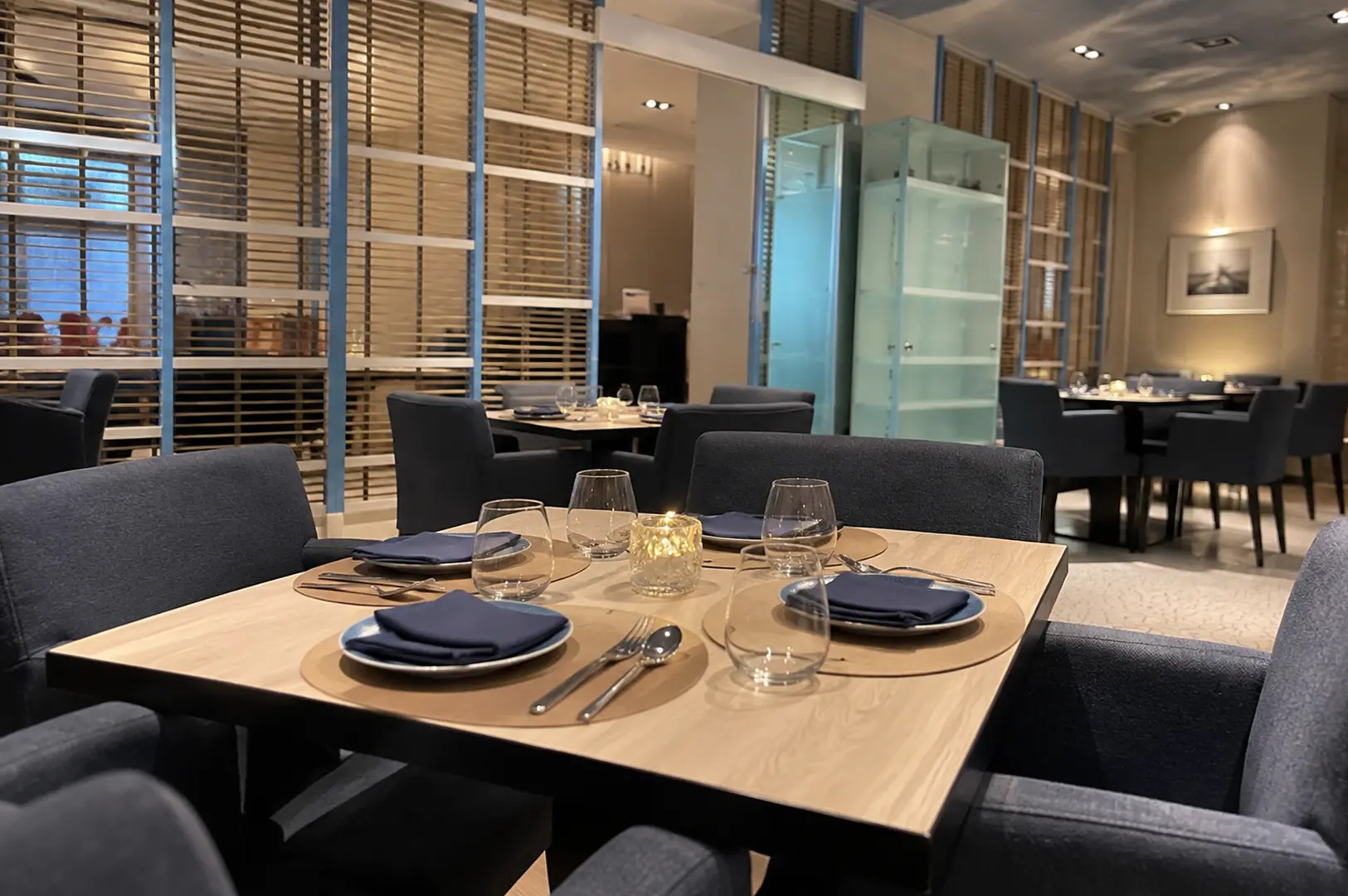Elegant restaurant dining room with neatly set wooden tables, navy chairs, glassware, and warm ambient lighting.