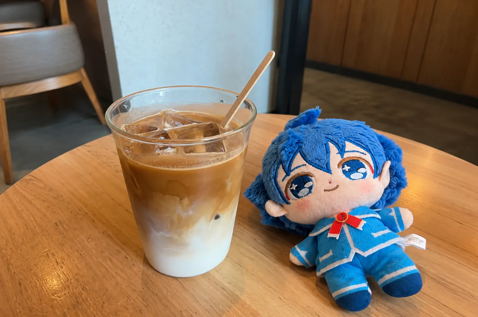 Iced latte beside a blue-haired plush doll on a wooden café table.