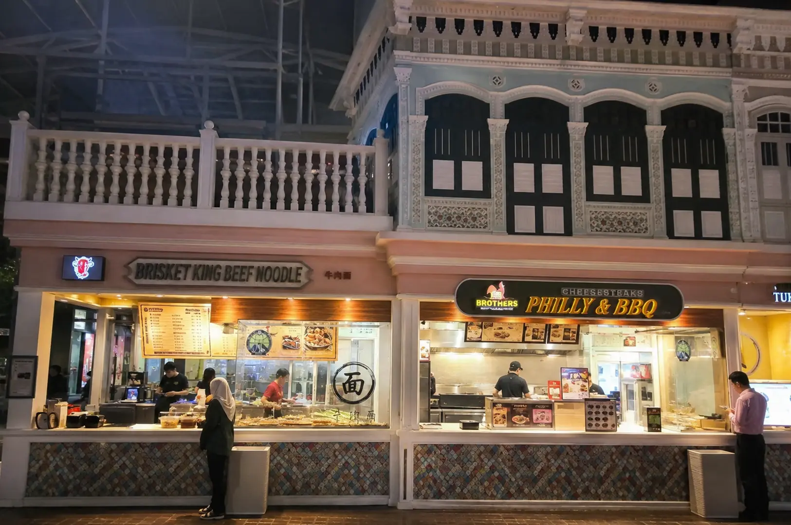 Night view of two street food stalls serving beef noodles and Philly BBQ sandwiches in a heritage-style building.