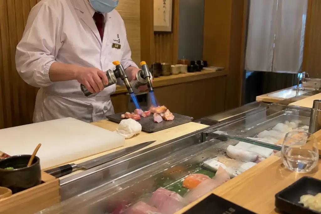 Japanese chef using two blowtorches to sear pieces of fish behind the sushi counter at Tatsuya