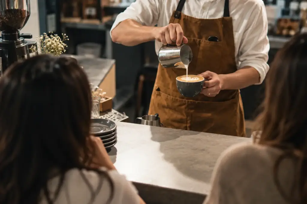 Barista pouring steamed milk into a coffee while customers watch at the counter.