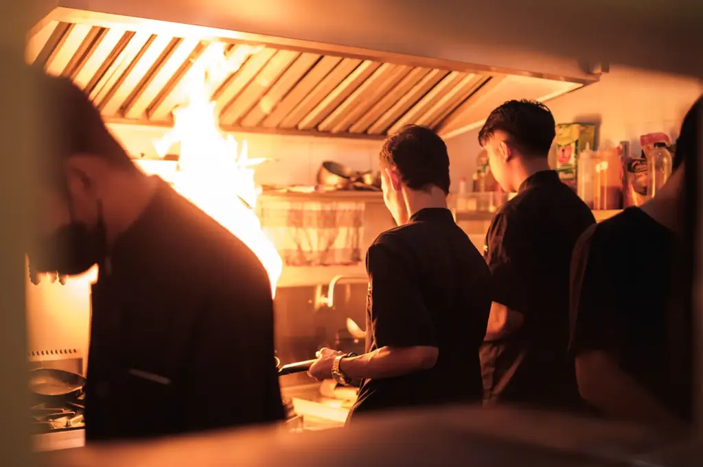 Restaurant kitchen staff working together around a busy cooking station with flames rising from a pan.