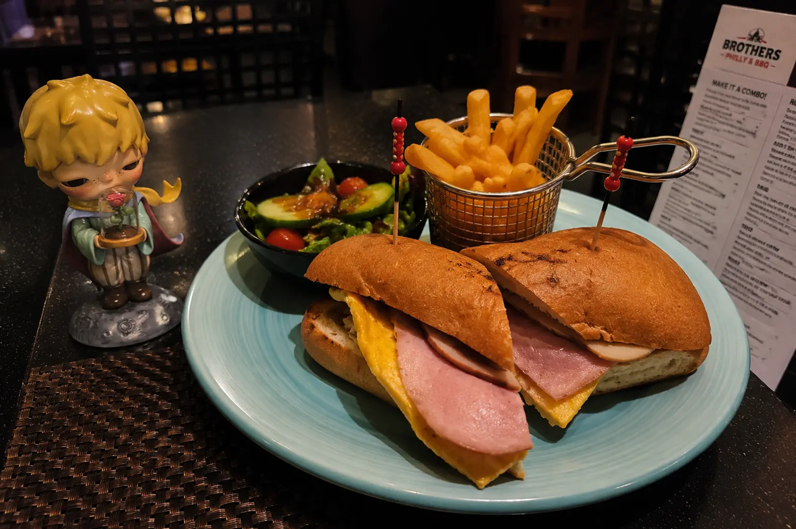 Ham and egg sandwich with fries and salad served on a plate beside a small decorative figurine.