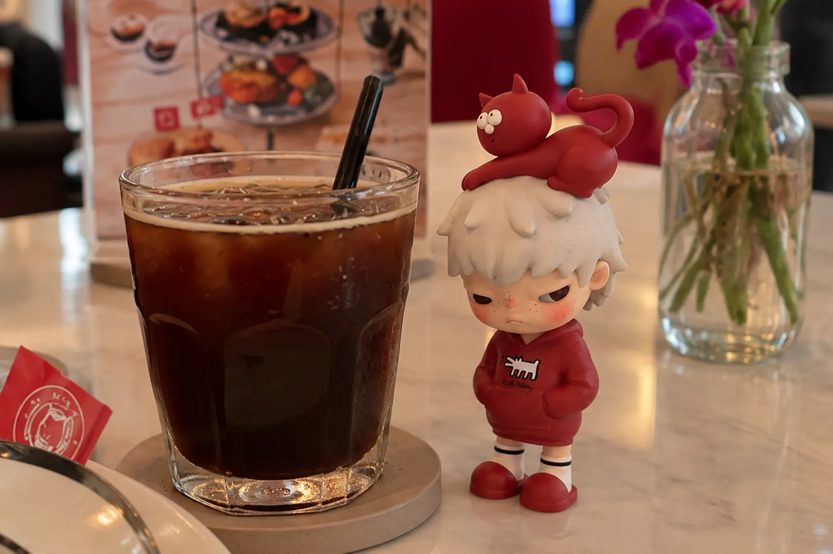 Iced coffee beside a red-and-white character figurine with a red cat on its head, displayed on a café table.