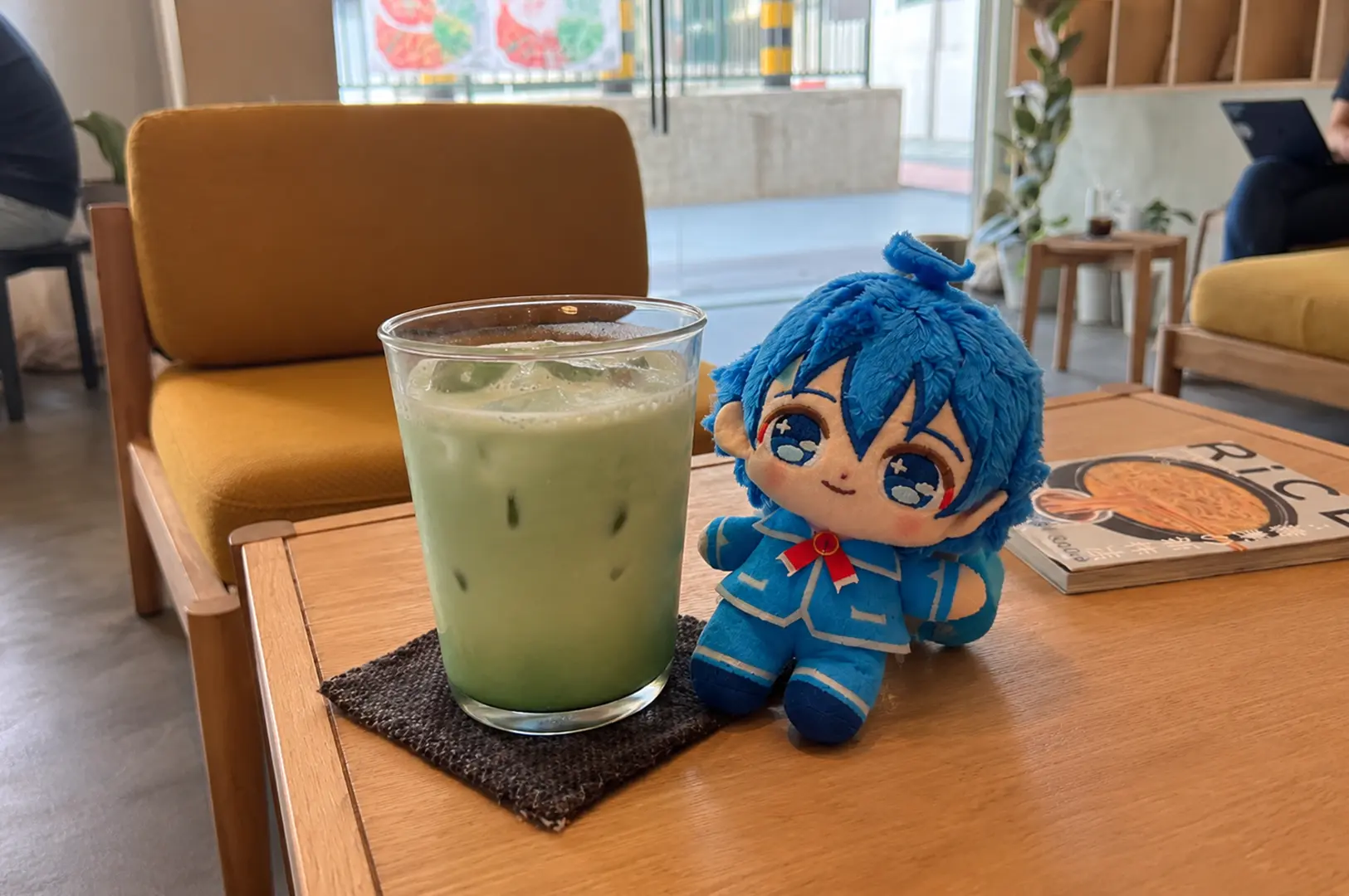 Iced matcha latte beside a blue-haired plush doll in a cozy café.