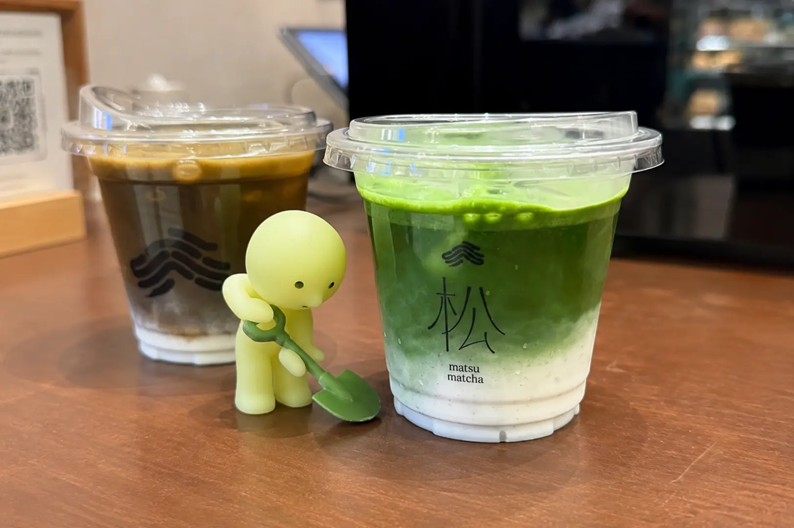Iced matcha latte and iced coffee beside a small green figurine.