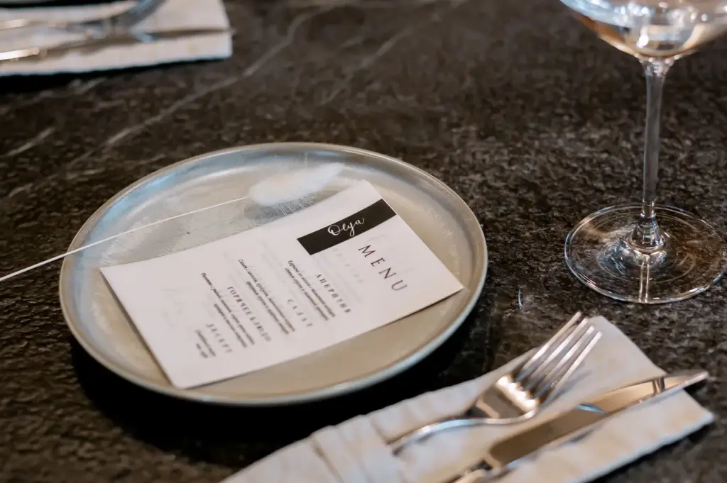 A restaurant menu resting on a plate beside polished cutlery and a wine glass.