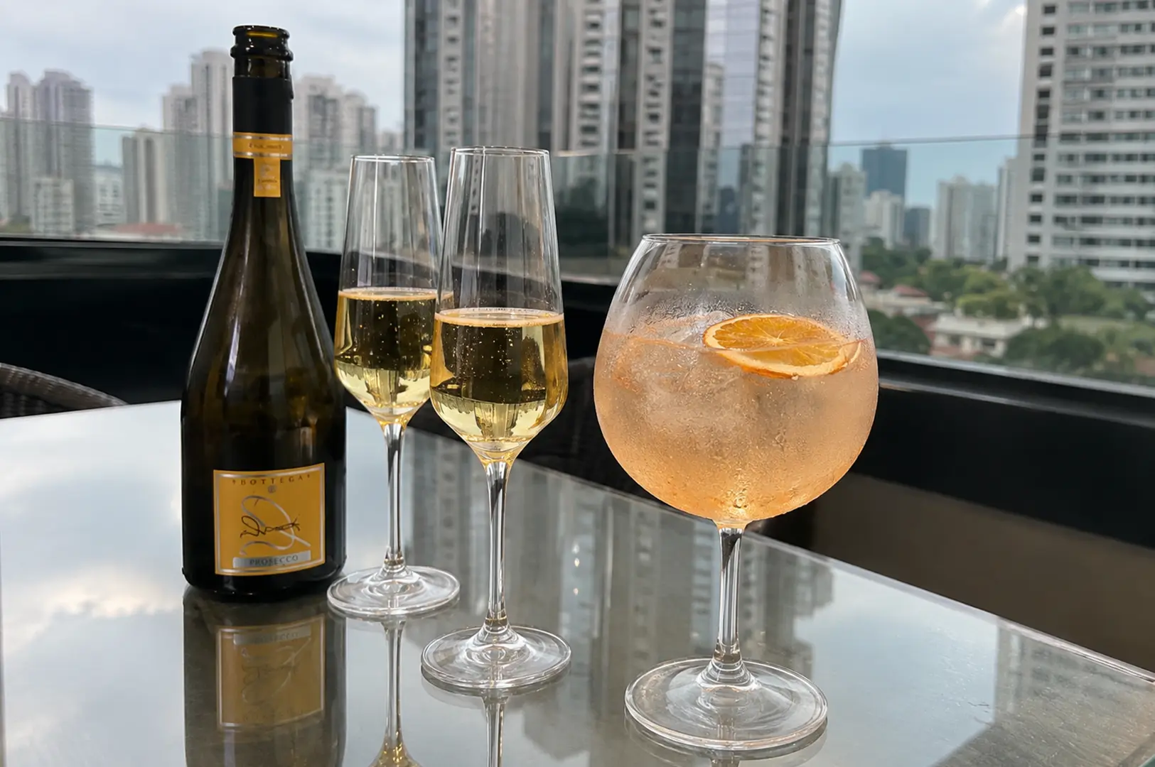 Bottle of Prosecco with two sparkling wine glasses and a citrus cocktail overlooking a city skyline.