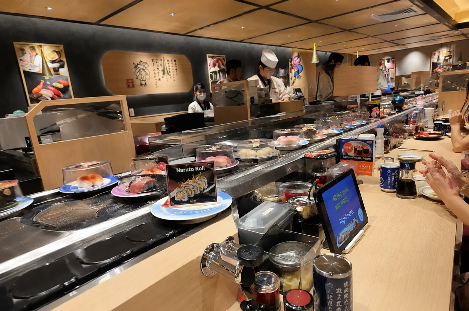 Modern conveyor belt sushi restaurant with chefs preparing dishes behind the sushi counter.
