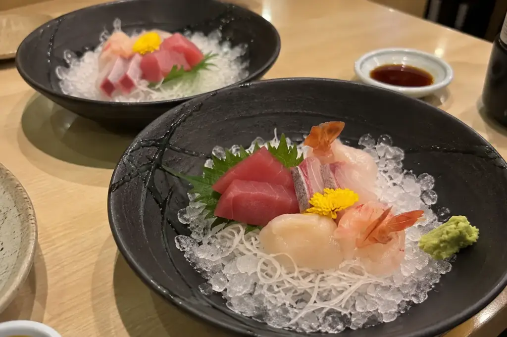 Assorted sashimi served on a black ceramic plate over crushed ice with shiso leaves, shrimp, tuna, and scallop.