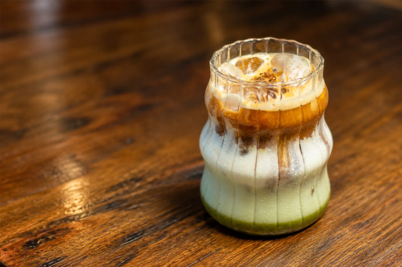 A close-up iced coffee served in a decorative glass on a warm wooden café table.