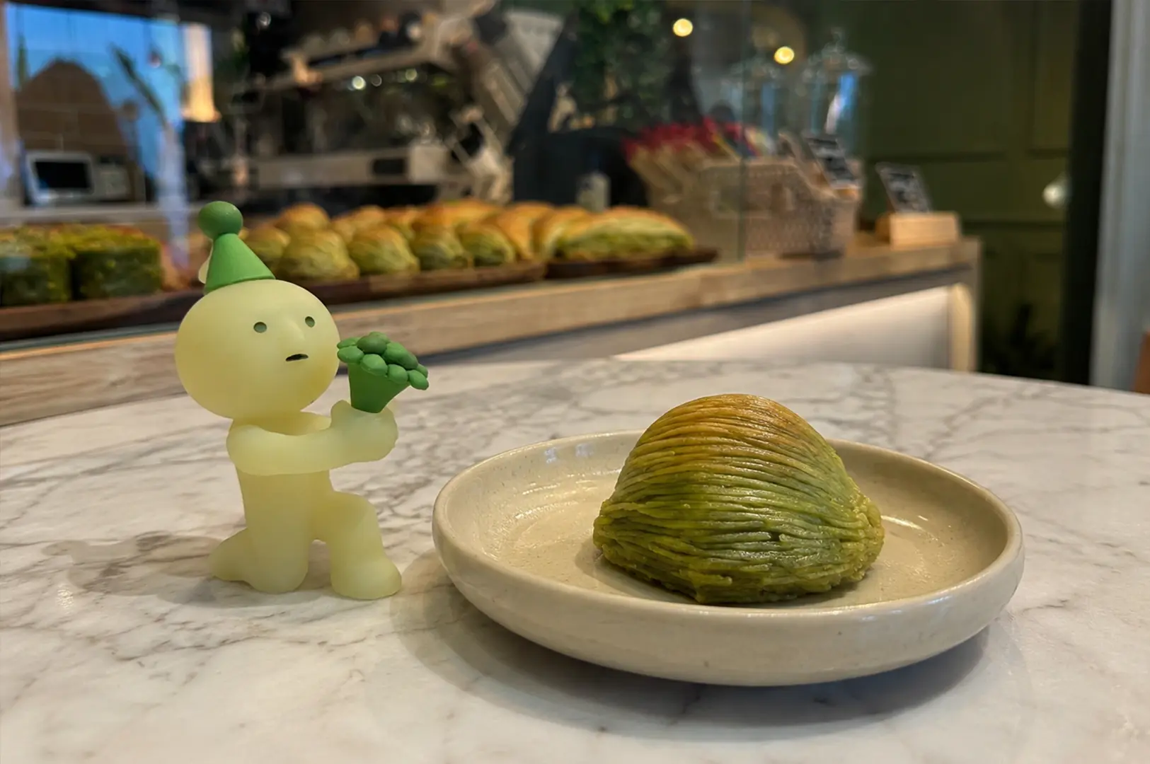 Smiski figurine beside a layered green pistachio dessert on a marble café table with a pastry display in the background.