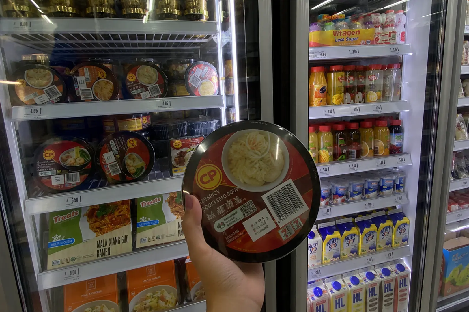 Shopper holding a ready-to-eat ramen bowl in front of a convenience store freezer filled with frozen meals and chilled drinks, capturing the modern grab-and-go food trend.