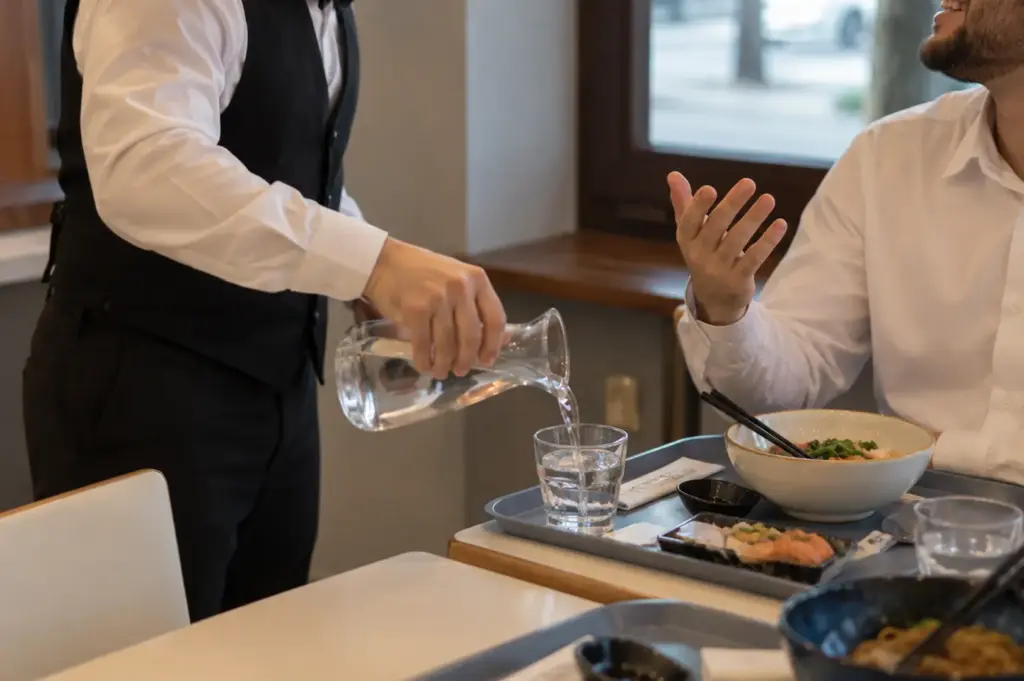 A waiter pouring water into a glass while a diner sits at the table with Japanese dishes.