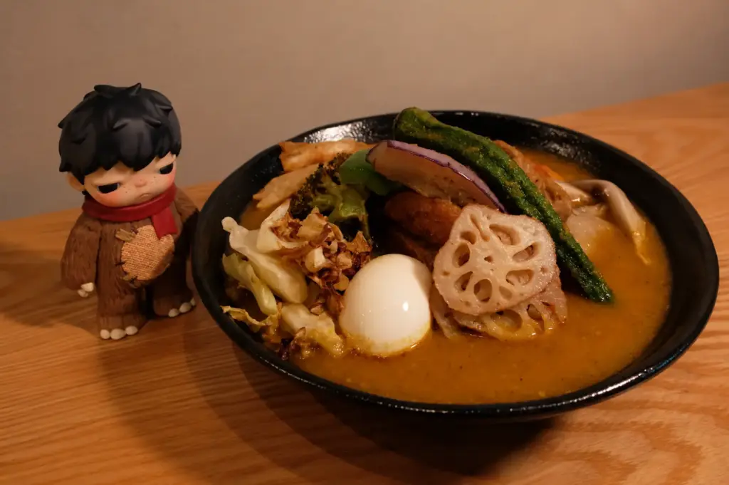 A colorful vegetable soup curry bowl with lotus root, egg, and a Hirono figurine at Soup Curry by Kisetsu.