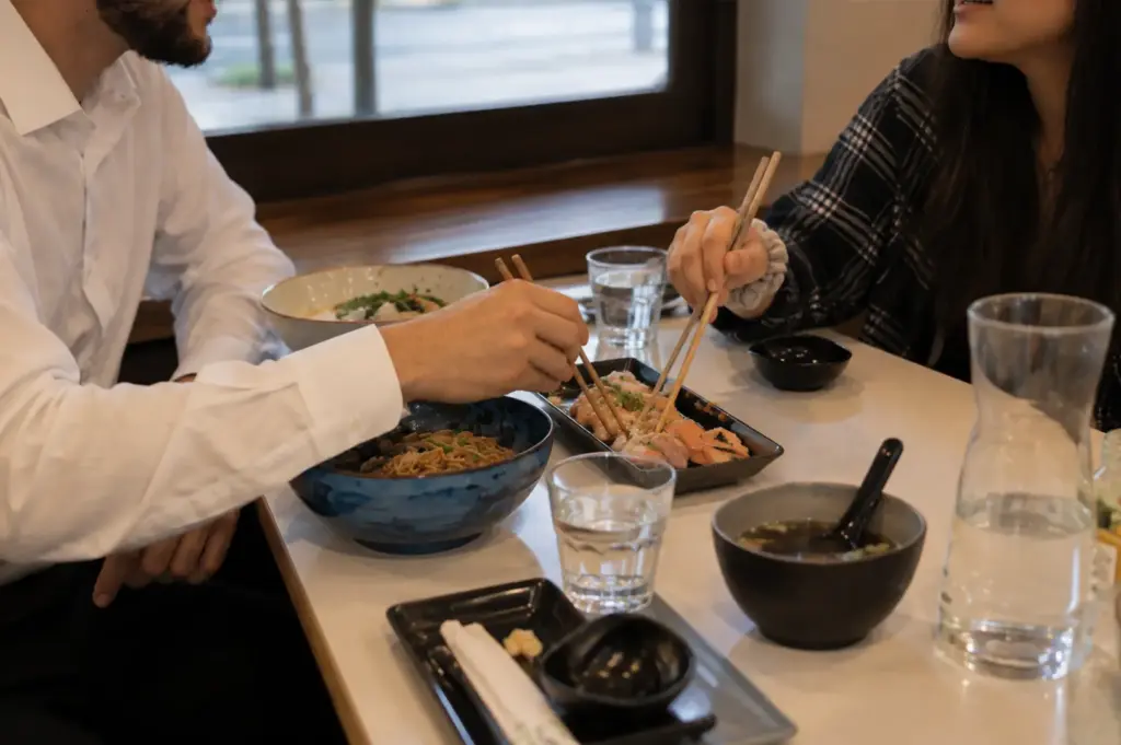 Two diners reaching for shared sushi with chopsticks while enjoying a casual restaurant meal.