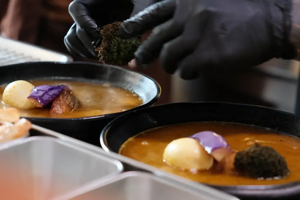 A chef wearing black gloves prepares soup curry bowls with vegetables and eggs at Soup Curry by Kisetsu.