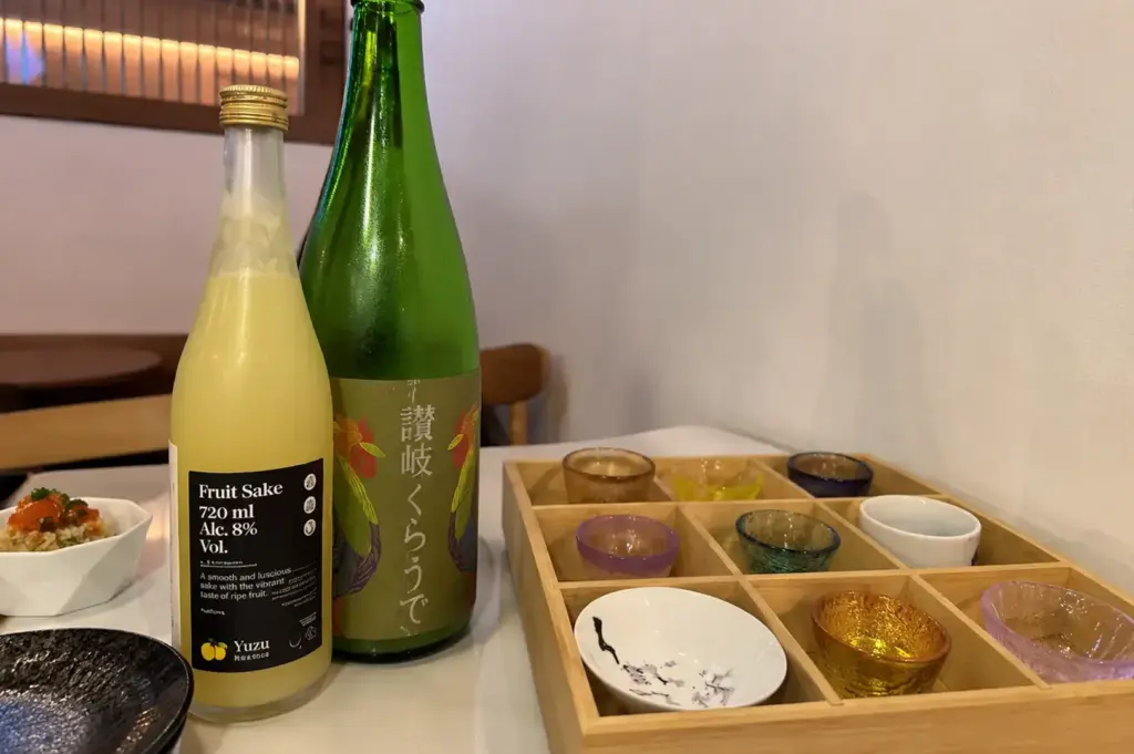 Japanese sake bottles and colorful tasting cups arranged on a table beside small dishes.