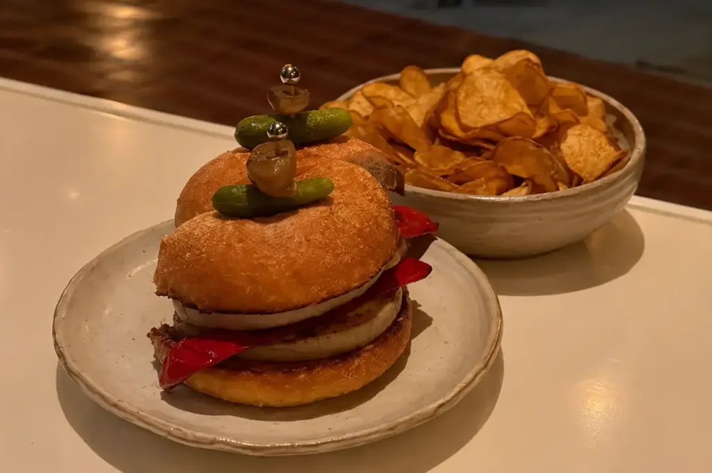 Gourmet sandwich with layered fillings and pickles, served with a bowl of crispy potato chips.